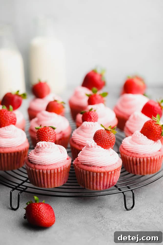 Many red cupcakes with pink frosting and whole strawberries on top, arranged on a round cooling rack.