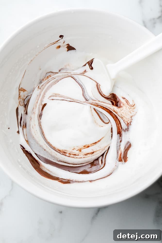 A white spatula gently folding rich melted chocolate into a fluffy white aquafaba mixture in a white mixing bowl.