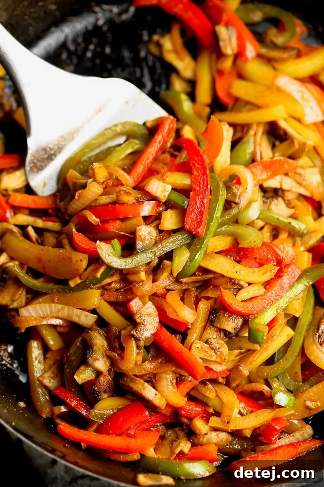 Close-up view of a spatula expertly mixing vibrant sliced bell peppers, onions, and mushrooms in a sizzling skillet, showing the cooking process of vegetarian fajitas.