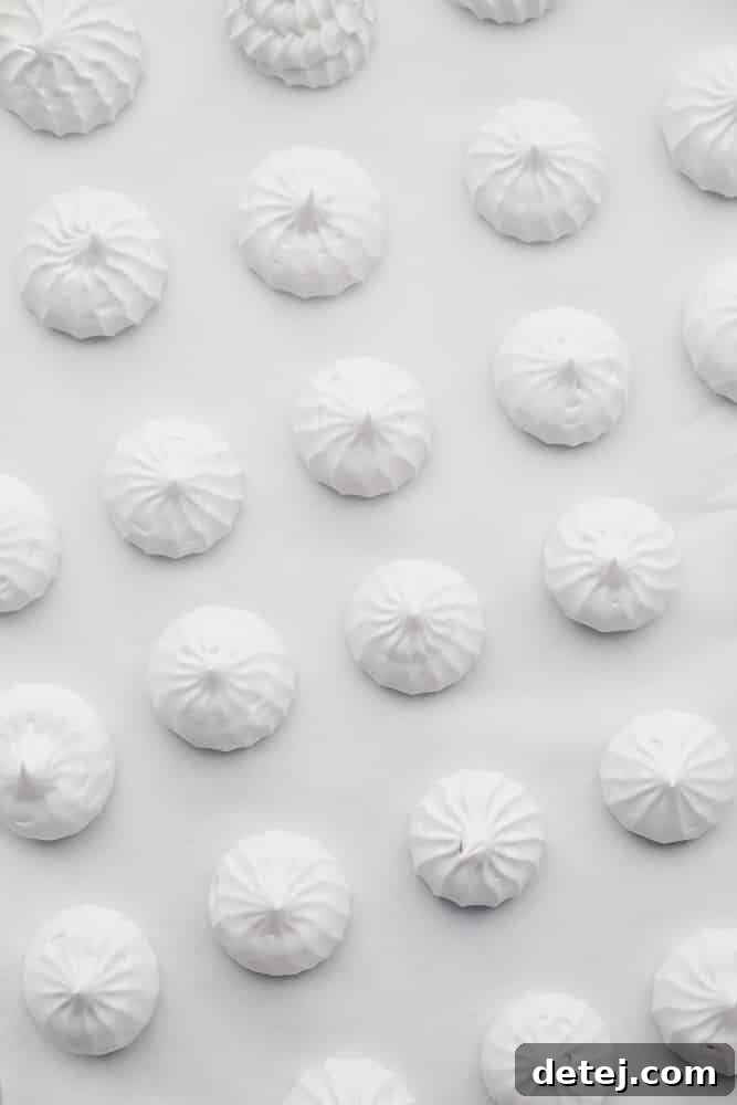 White meringue cookies cooling on parchment paper on a baking sheet, showcasing their even shapes.