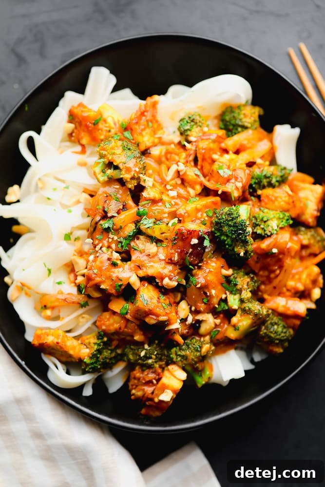 orange tempeh stir fry on top of white noodles in a black bowl