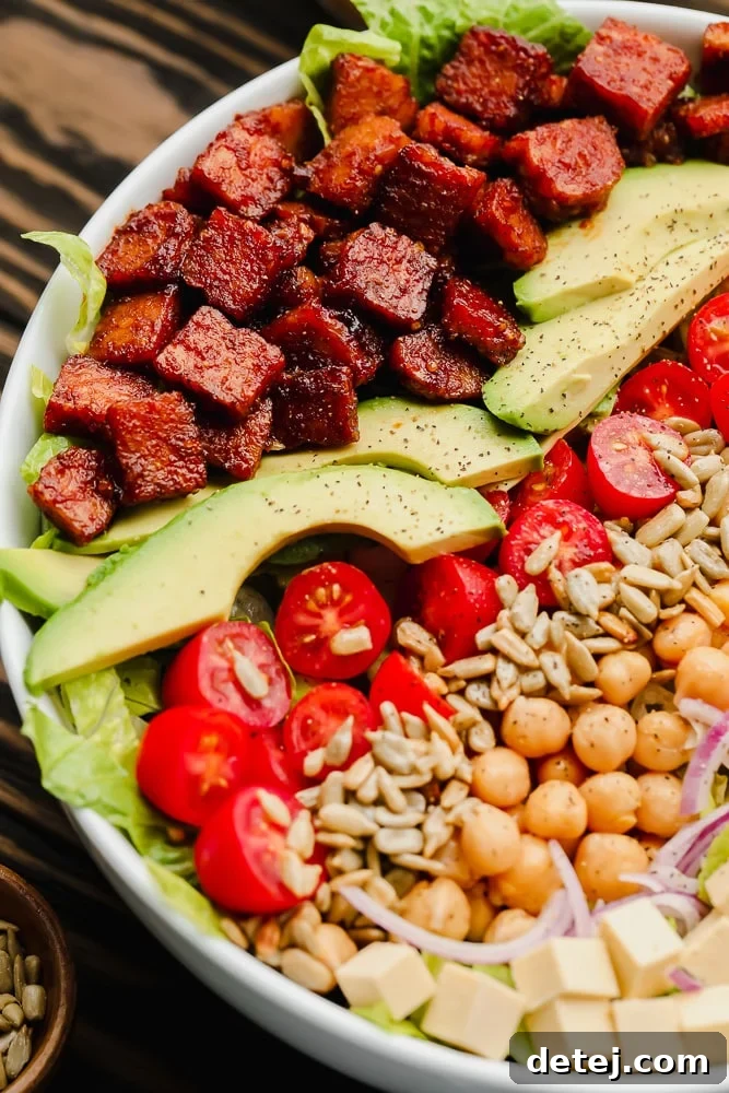 close up of lines of vegan bacon, avocado, tomatoes, seeds, and beans on top of lettuce in a white bowl
