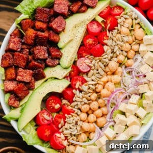 close up of lines of vegan bacon, avocado, tomatoes, seeds, and beans on top of lettuce in a white bowl