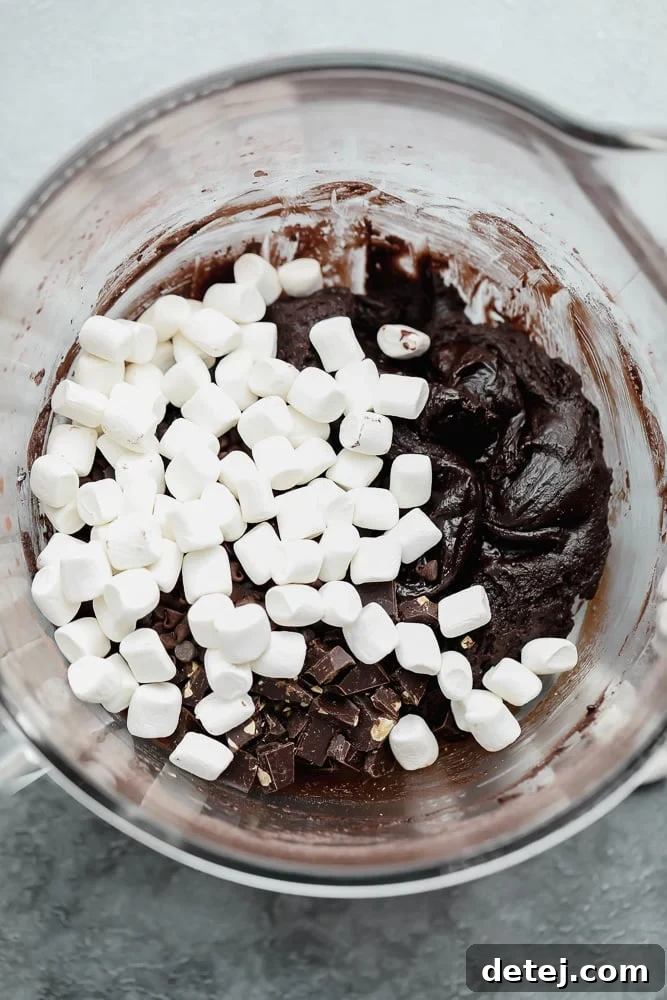 A large white bowl filled with rich, dark chocolate cookie batter, generously mixed with white mini vegan marshmallows, ready for baking.