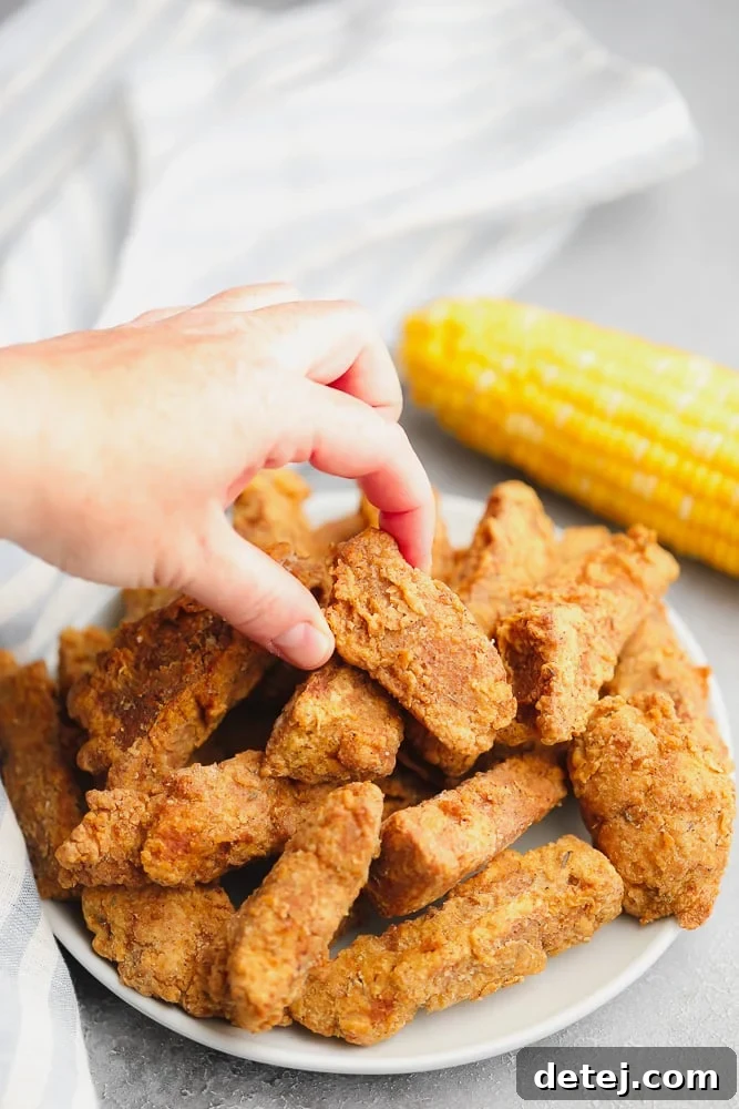 A woman's hand reaching for a piece of golden-brown fried vegan chicken from a generous pile on a white plate, highlighting its tempting appearance.