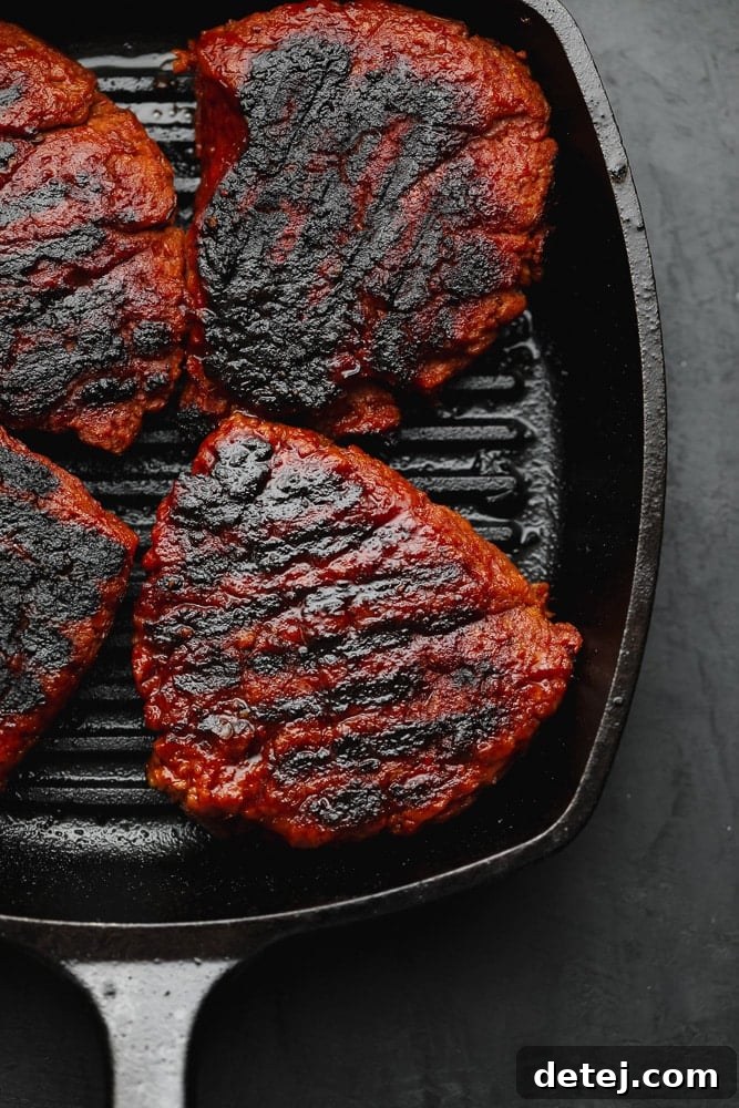 Four perfectly charred vegan steak patties resting in a black cast iron grill pan, showcasing their smoky texture and appealing color.