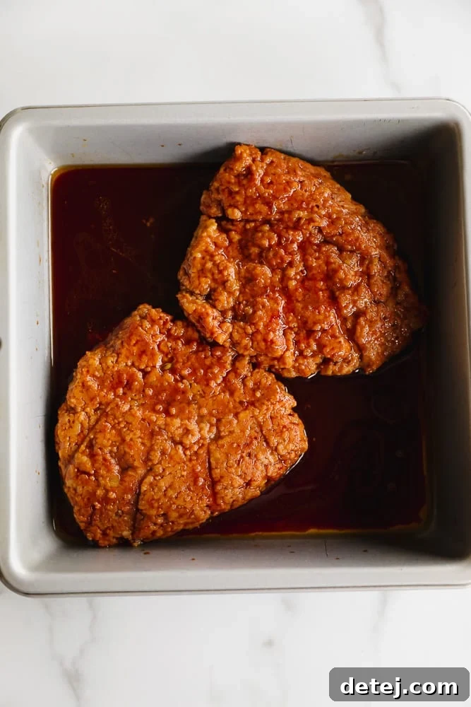 Two vibrant orange seitan patties generously marinating in a rich, dark brown liquid in a shallow dish, ready to absorb delicious flavors.