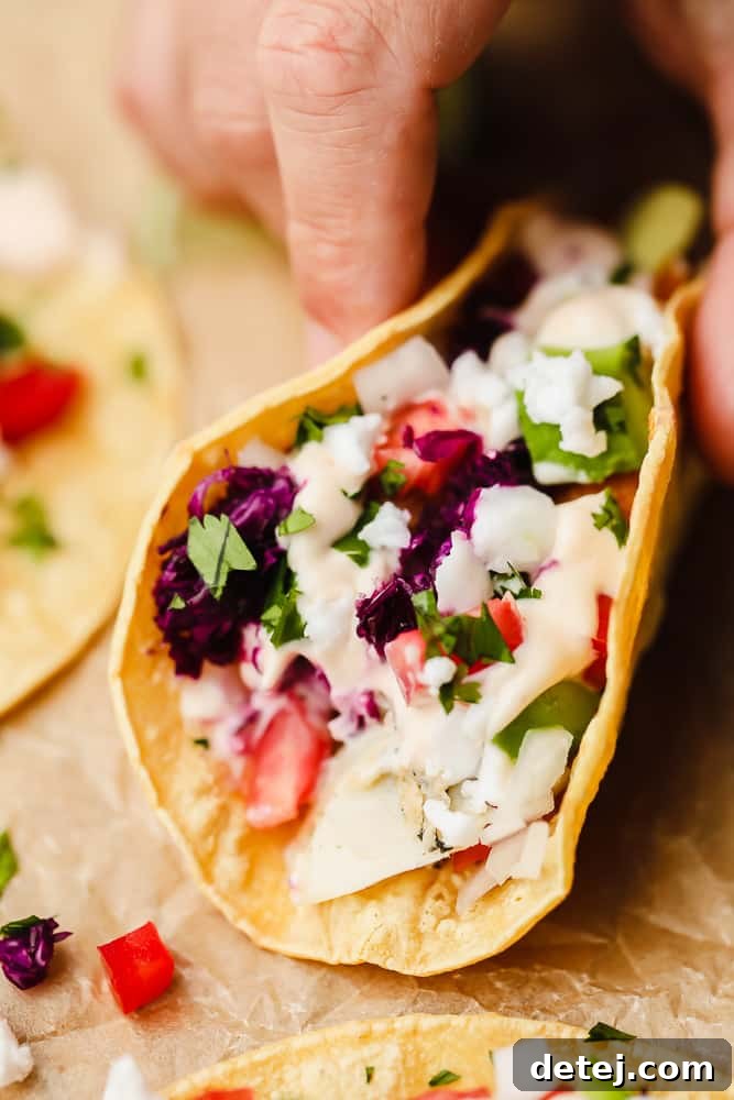A hand reaching for a vibrant corn tortilla filled with creamy white sauce, diced red tomatoes, and shredded purple cabbage, highlighting the fresh ingredients.