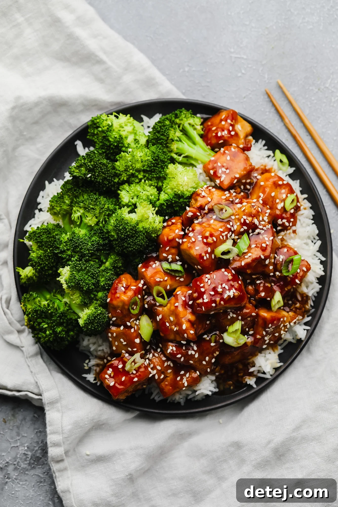 A black plate featuring golden-crisp teriyaki tofu chunks, alongside a serving of white rice and bright green broccoli florets, set against a subtle grey background.