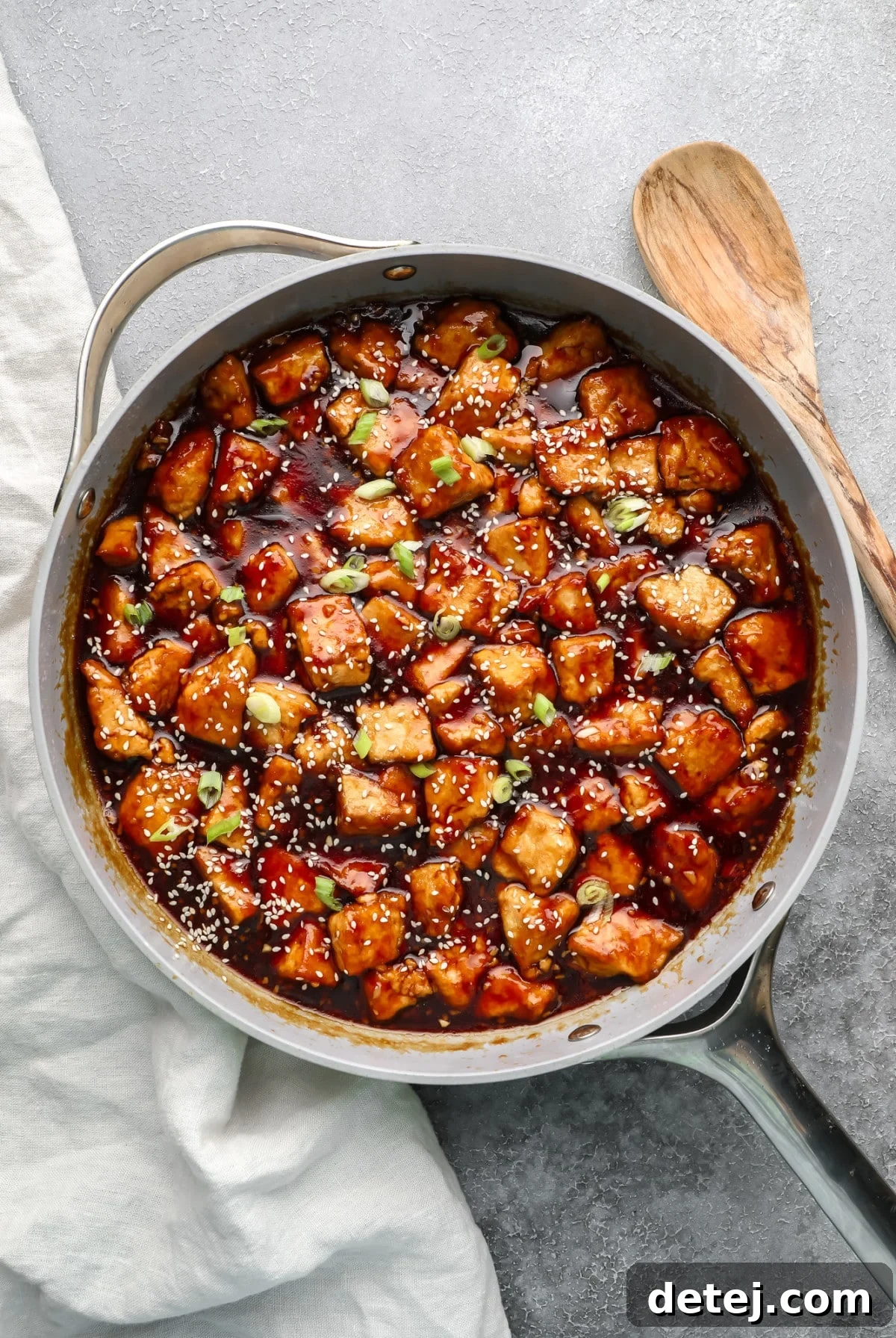 A large grey pan filled with glossy teriyaki tofu chunks, generously garnished with sesame seeds and chopped green onions.