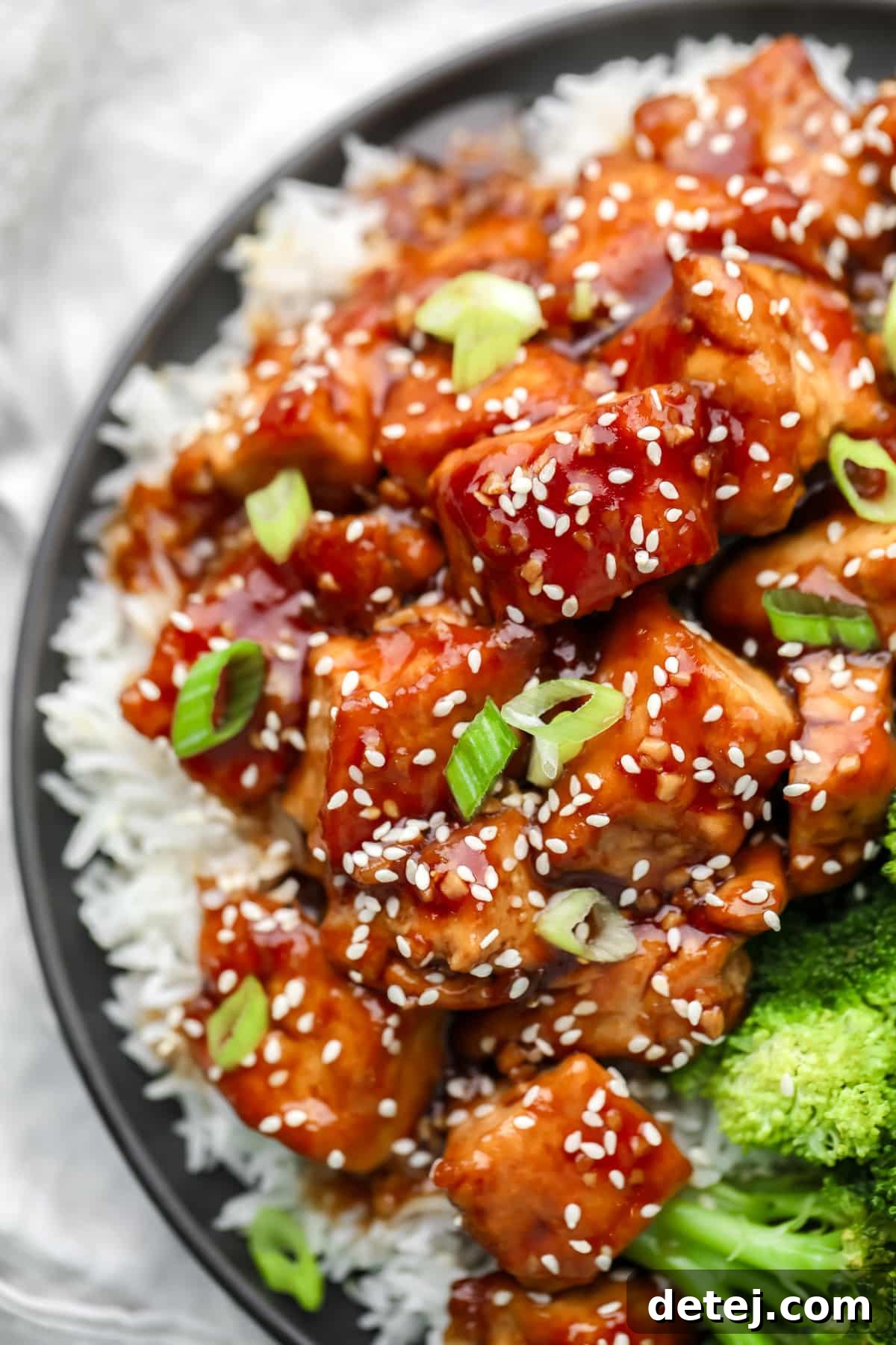 A plate featuring a generous portion of white rice topped with saucy brown teriyaki tofu and sprinkled with sesame seeds, presented against a clean background.