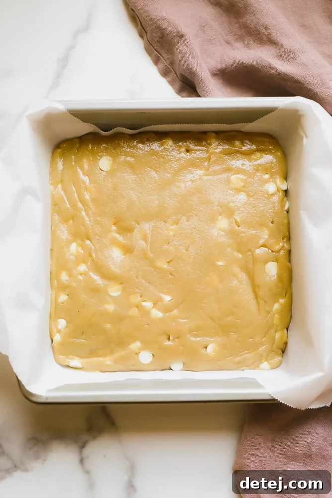 Light brown blondie batter evenly spread in a parchment-lined baking dish, ready for the oven