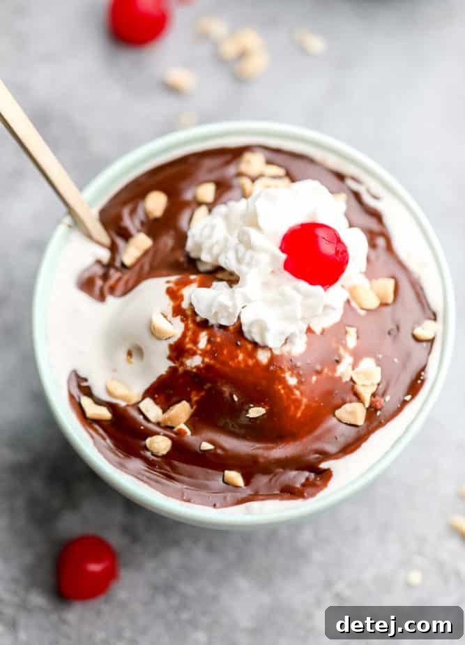 Teal bowl of vegan vanilla ice cream generously drizzled with rich vegan hot fudge, topped with fluffy coconut whipped cream and a bright red cherry.
