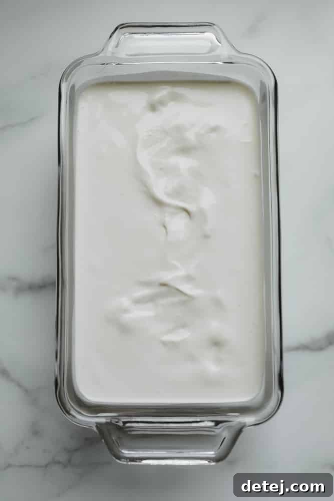 A loaf pan filled with a smooth, white mixture, covered with plastic wrap, ready for freezing into vegan ice cream.
