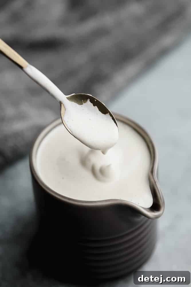 A golden spoon scooping creamy vegan heavy cream from a small grey pitcher, with a soft focus background.