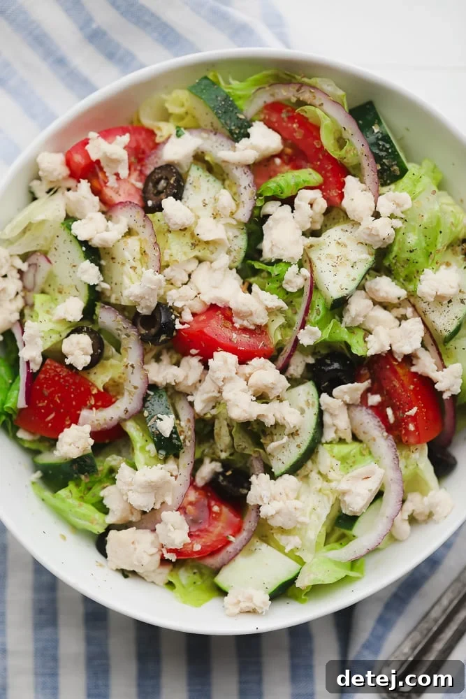 a Greek salad in a white bowl topped with crumbled white vegan feta cheese and fresh herbs