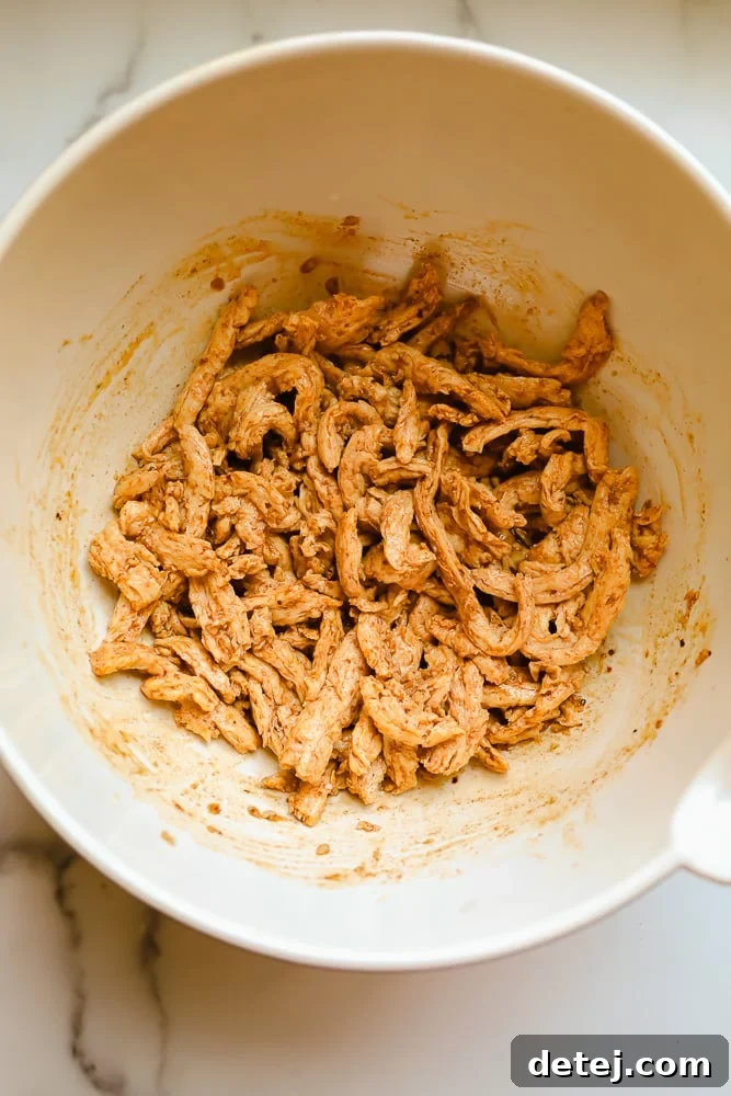 Rehydrated soy curls being mixed in a white bowl with a rich, orange-hued seasoning blend, ensuring every piece is coated in the aromatic marinade.