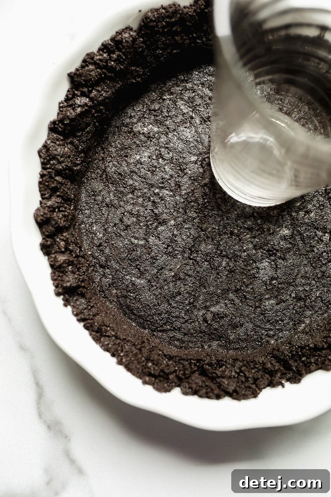 A hand using a water glass to firmly press a dark brown cookie crust mixture into a white pie pan, creating an even base.