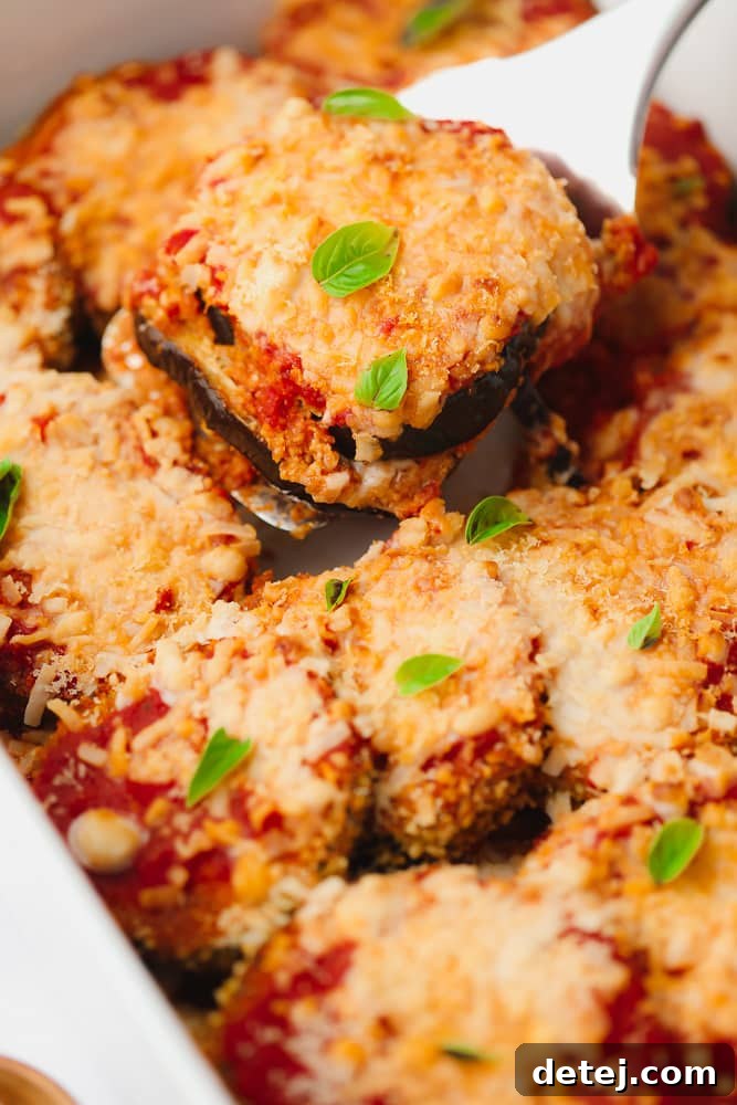 Spatula scooping a golden-brown slice of vegan eggplant parmesan from a white baking dish, showing layers of crispy eggplant, red marinara, and melted white cheese.