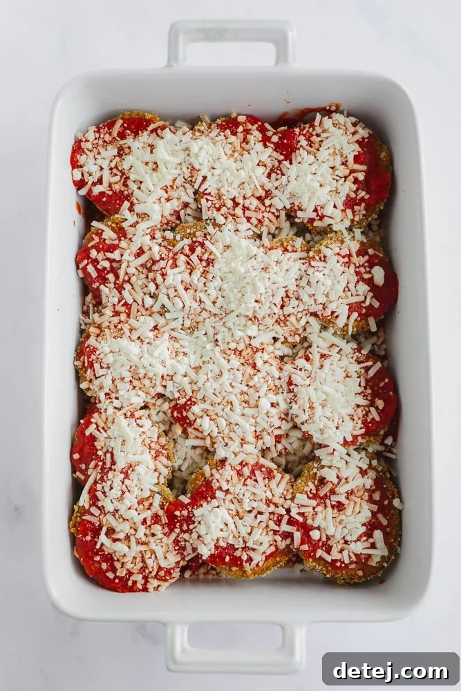 Layers of golden-baked eggplant slices, rich red tomato sauce, and perfectly melted white vegan cheese nestled in a rectangular white baking dish, ready for its final bake.