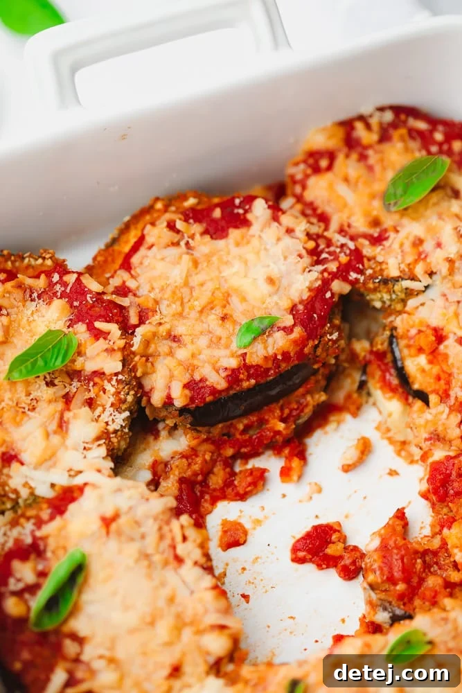 A large white baking dish filled with baked vegan eggplant parmesan, generously topped with melted vegan cheese, vibrant marinara sauce, and fresh green herbs.