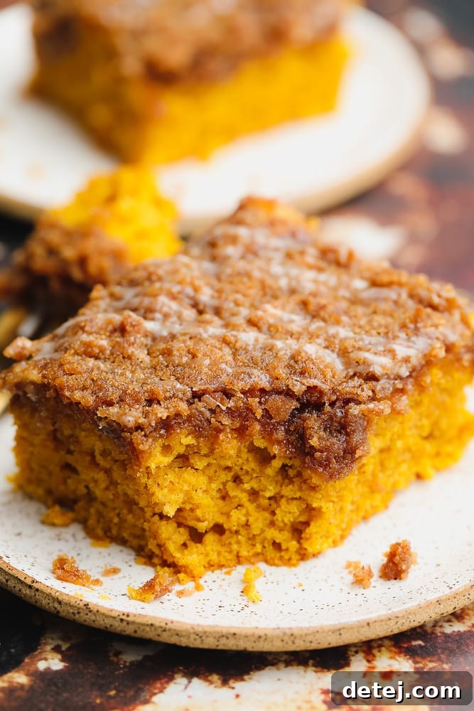 Close-up on a square slice of moist, orange vegan pumpkin coffee cake on a white plate, showcasing the streusel topping and a drizzle of glaze.