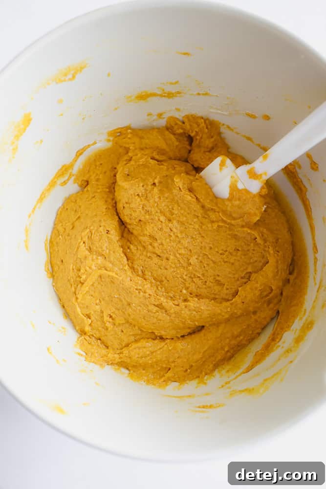 White spatula mixing a vibrant orange pumpkin cake batter in a white bowl, showing the creamy texture.