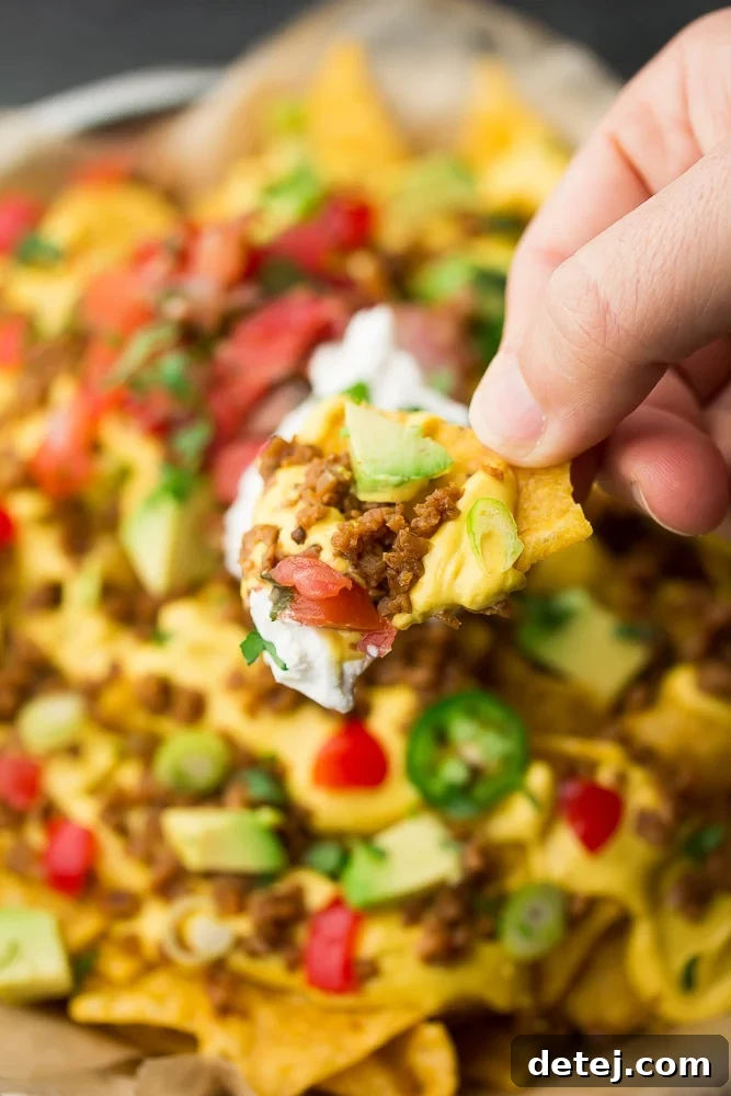 A hand reaching into a pile of fully loaded vegan nachos, picking up a chip laden with savory toppings.