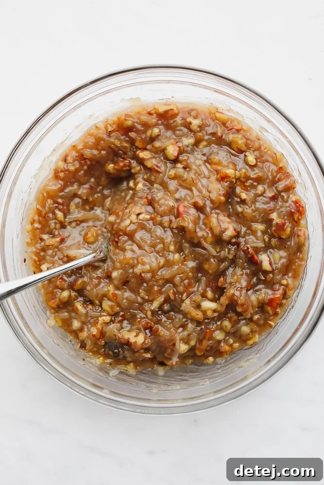 A spoon stirring a thick, luscious caramel sauce with pecans and coconut in a glass bowl after chilling.