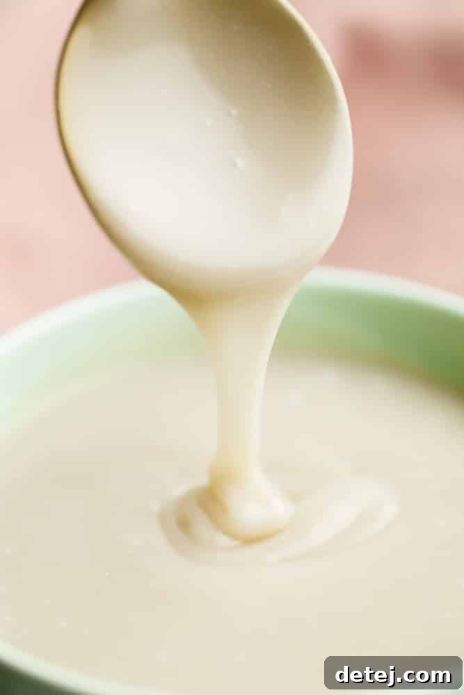 A spoon drizzling a creamy white liquid into a green bowl, showcasing the perfect consistency of homemade vegan condensed milk.