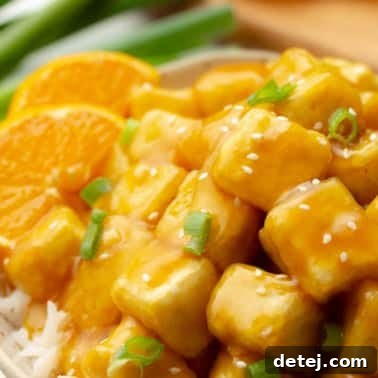 close up on orange tofu with orange slices on the side and sliced green onions on top.
