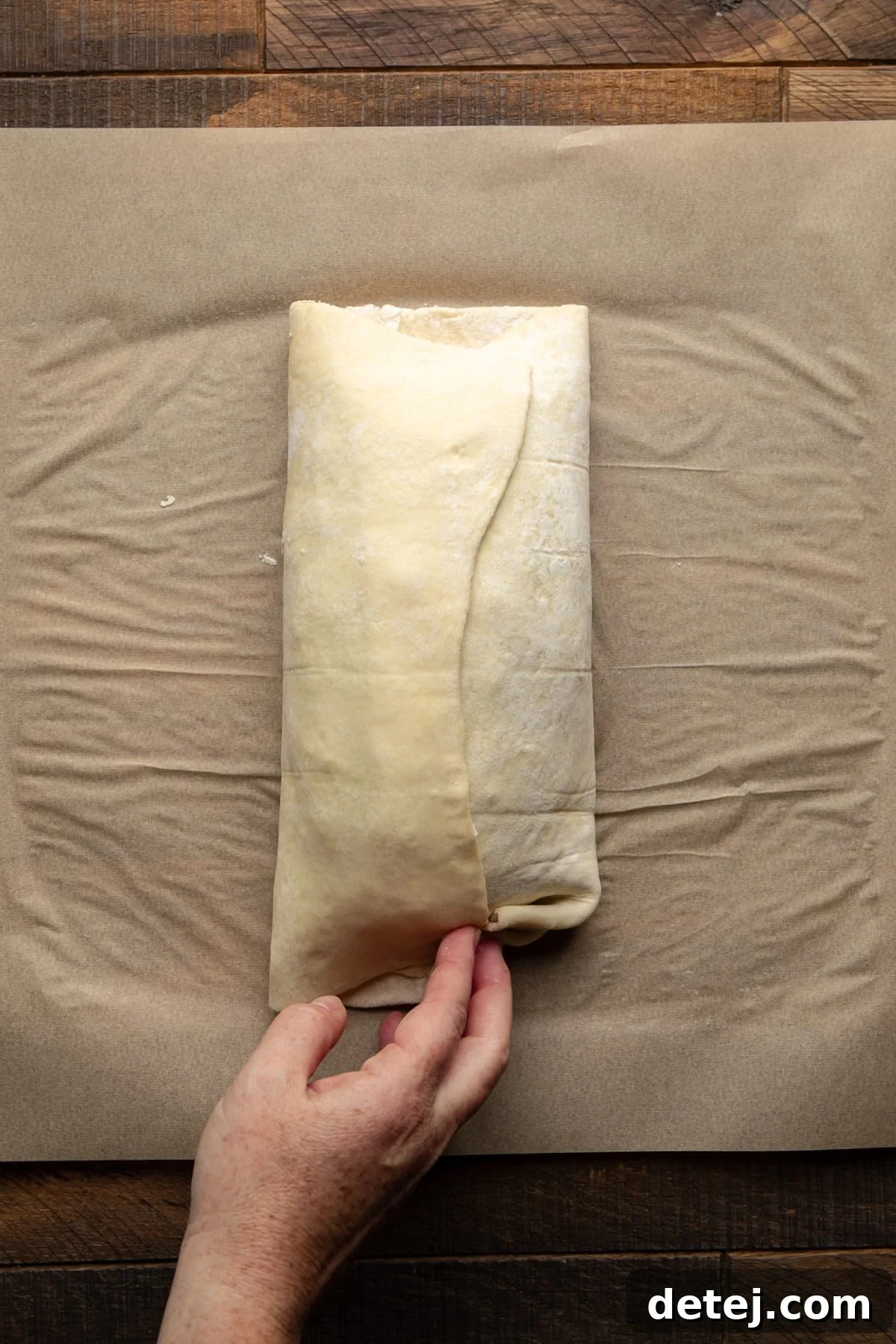Hands carefully sealing the edges of a pastry-wrapped vegan wellington on parchment paper.