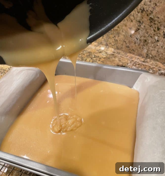 Gooey caramel being poured smoothly over a baked shortbread crust in a parchment-lined baking dish.