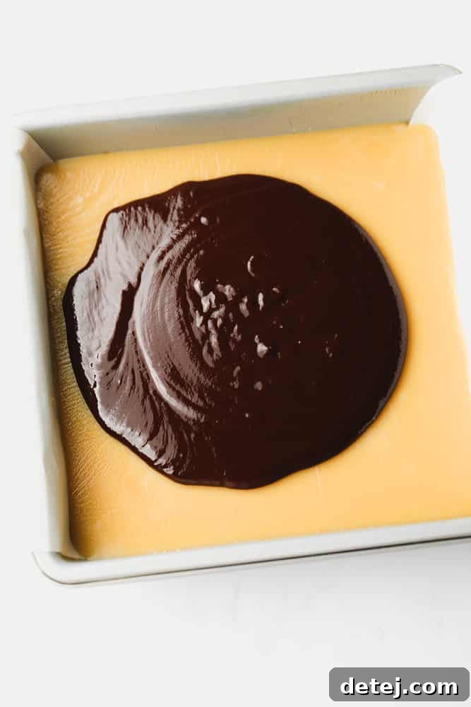 Pouring a smooth, dark melted chocolate ganache over a golden caramel layer in a metal baking dish.
