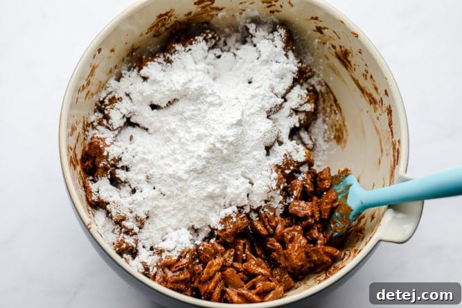 A bowl of chocolate-coated rice Chex cereal with powdered sugar being added, illustrating the final step of creating the classic muddy buddies coating.
