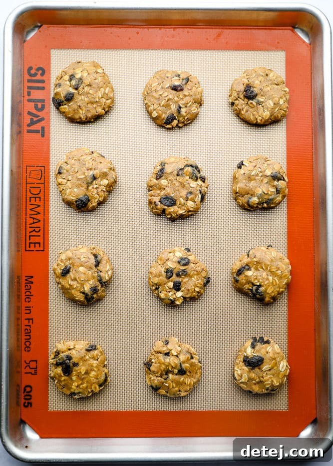 Unbaked vegan oatmeal raisin cookie dough balls neatly arranged on a silicone mat-lined baking tray, slightly flattened and spaced apart.