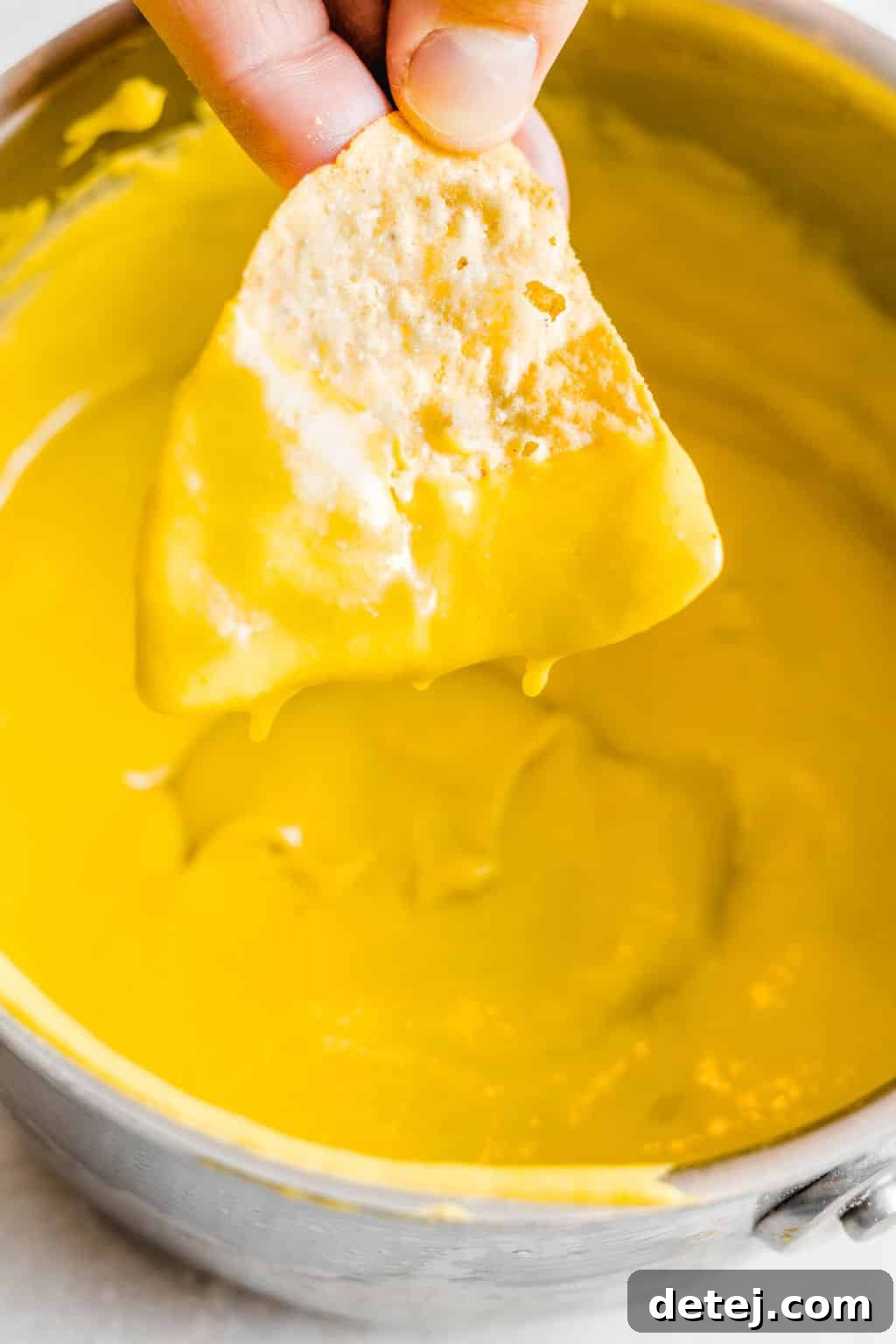 Woman's hand dipping a nacho chip into a pot filled with vibrant yellow vegan cheese sauce.