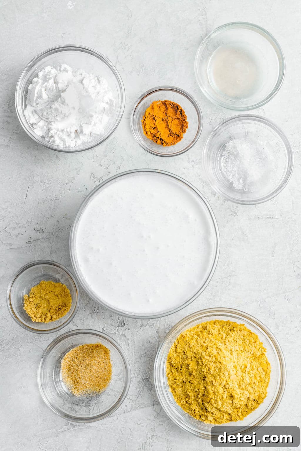 Individual glass bowls filled with various yellow seasonings, white powder, and plant milk, representing the simple ingredients for vegan cheese sauce.