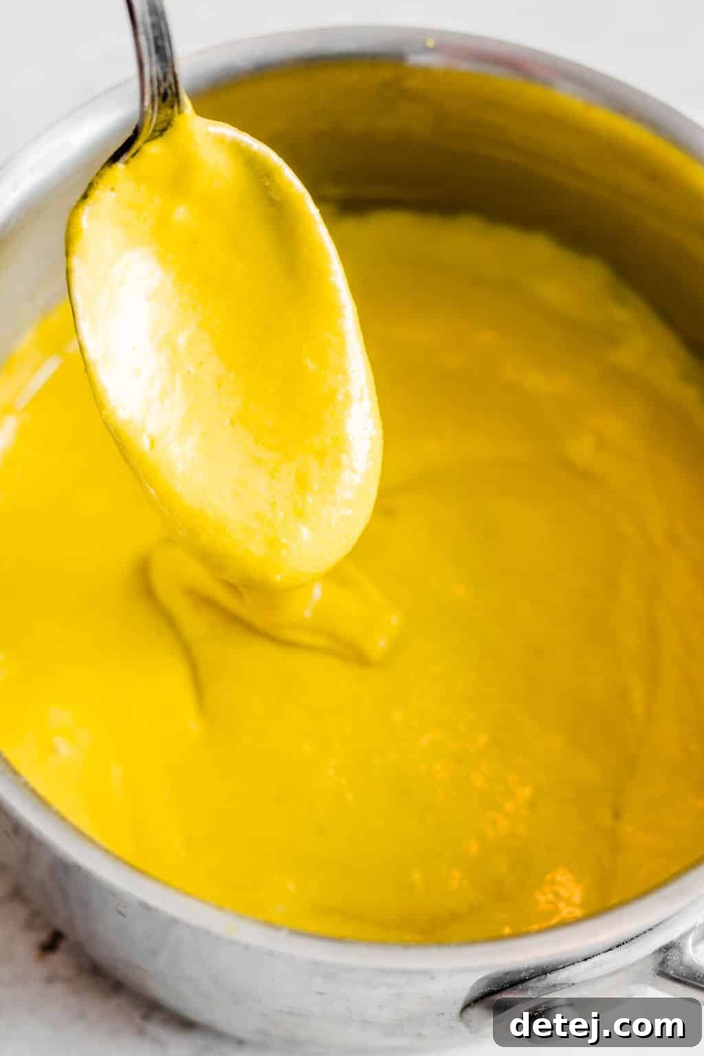 Close up on a spoon taking a scoop of warm, yellow vegan cheese sauce out of a metal pot, highlighting its creamy texture.