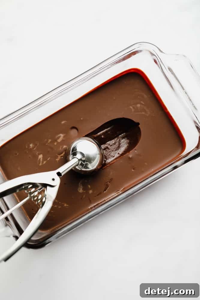 A metal ice cream scoop delicately scooping a portion of rich chocolate mixture from a glass dish, poised to form a truffle.