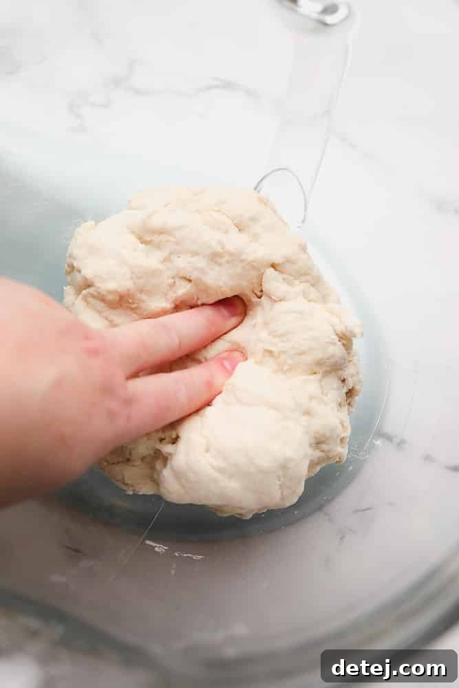 Fingers gently pressing into the soft dough within a glass mixing bowl to check for stickiness.