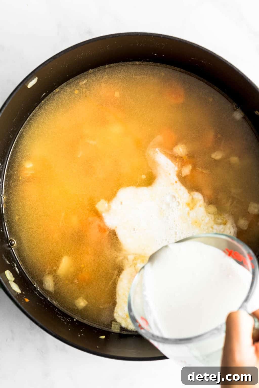 A woman's hand carefully pouring white vegan creamer into a black pot already filled with simmering broth and vegetables.