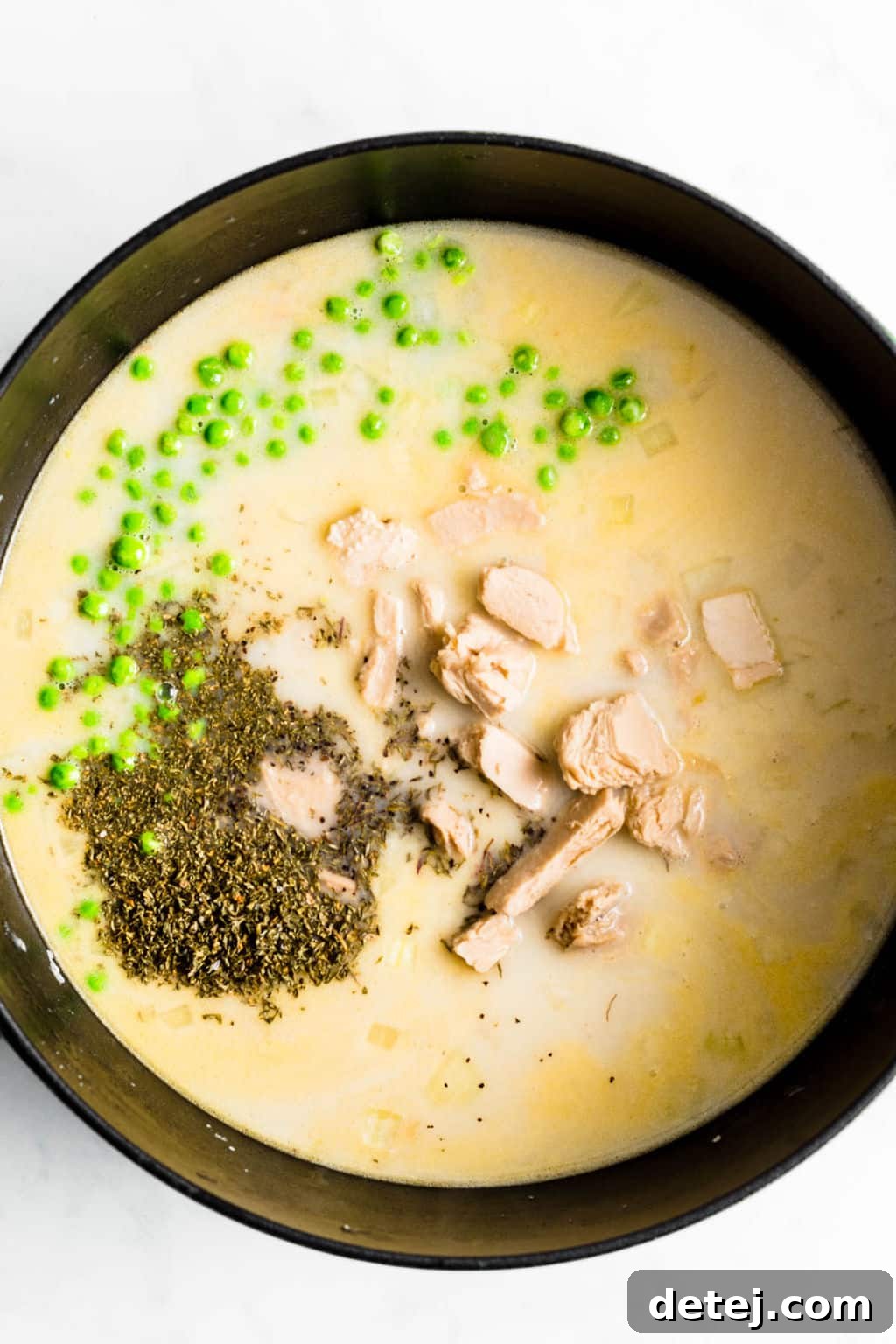 A large black pot filled with a creamy, savory vegan chicken soup, complete with plant-based chicken pieces, dried herbs, and vibrant green peas, simmering gently.