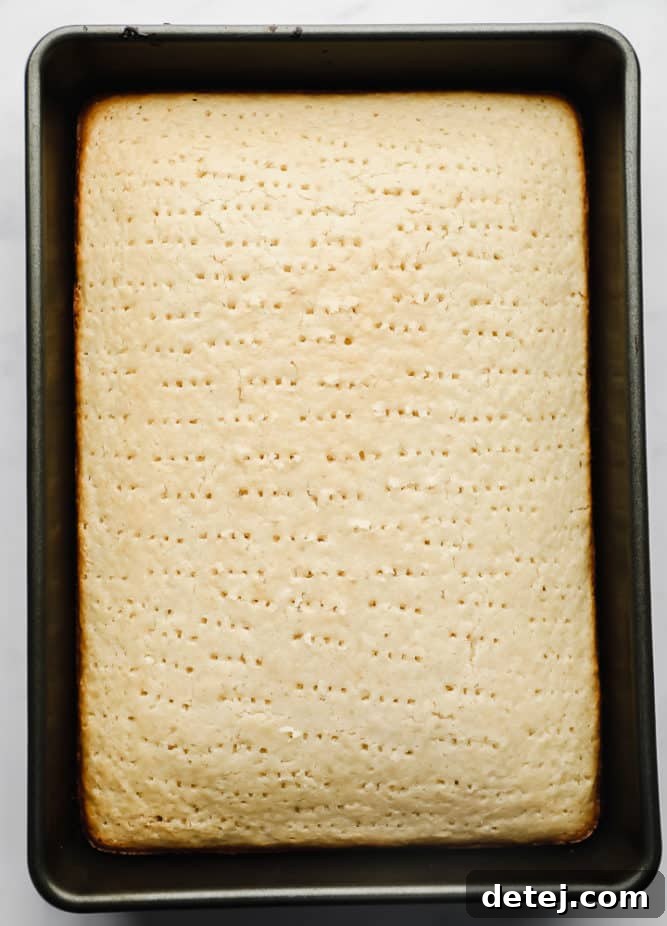 A vanilla cake in a baking pan, covered with numerous small holes poked into its surface, ready for the milk soak.