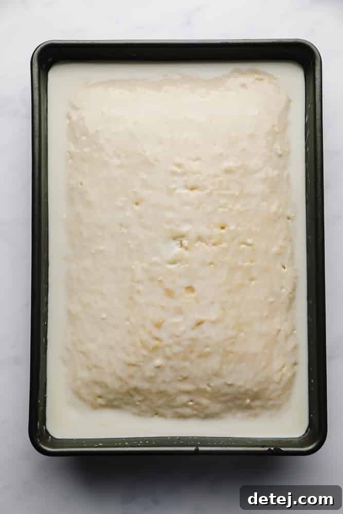 A vanilla cake in a pan with the rich, creamy milk mixture being poured generously over its surface, ready for soaking.