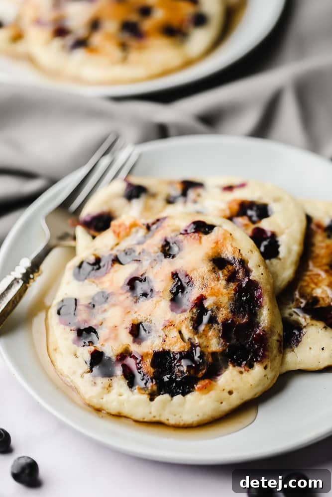 A stack of three golden-brown vegan blueberry pancakes, generously studded with fresh blueberries, on a pristine white plate, ready to be enjoyed.