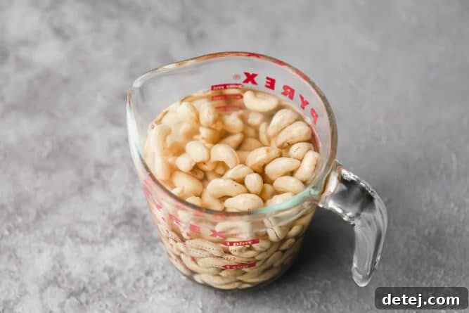 Raw cashews soaking in hot water inside a clear glass measuring cup, softening before blending.