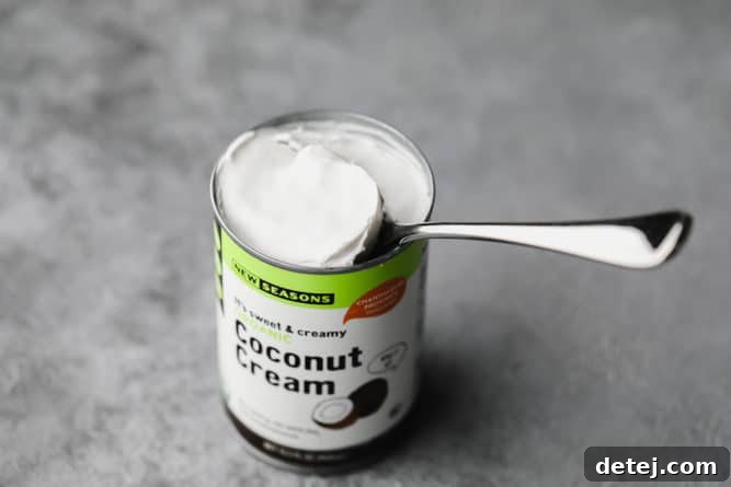 An open can of full-fat coconut cream with a spoon scooping out the thick white cream, illustrating how to separate it from the liquid.