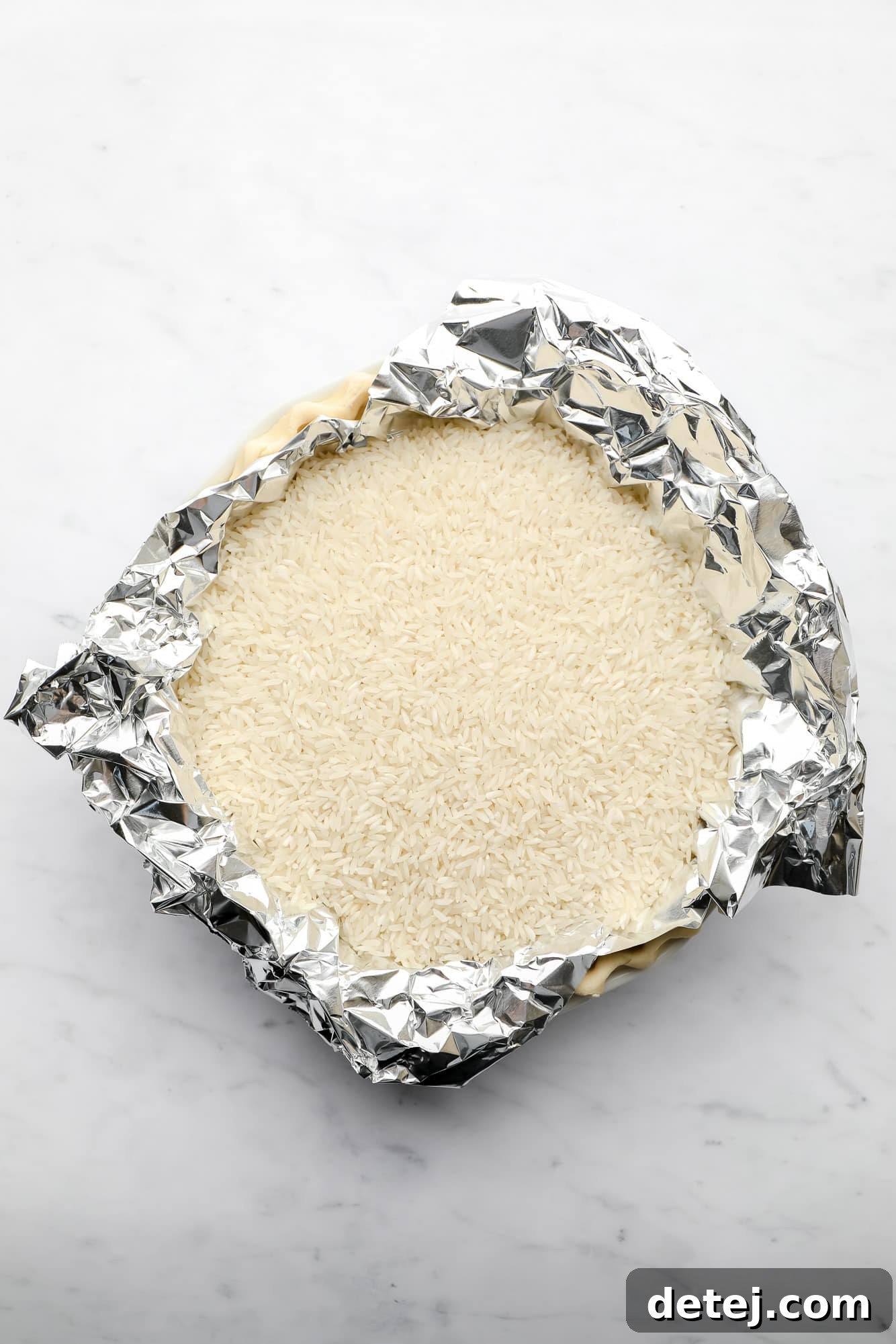 A pie crust baking in a pie plate, weighted down with a layer of tinfoil and white rice to prevent shrinking.