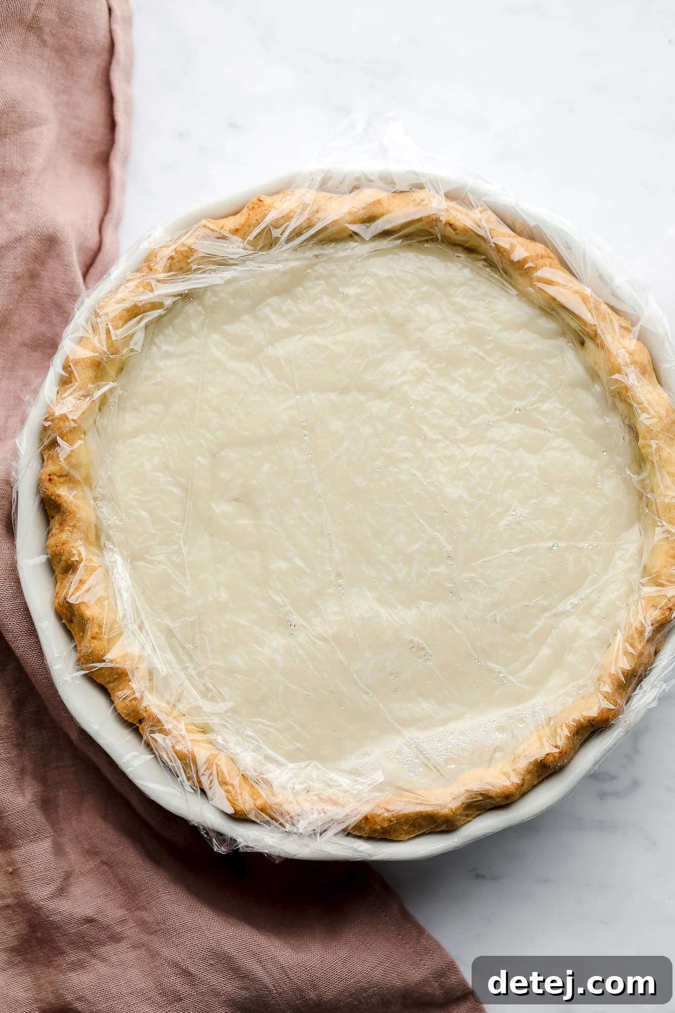 A smooth coconut custard filling inside a pie crust, covered with a layer of plastic wrap for chilling.